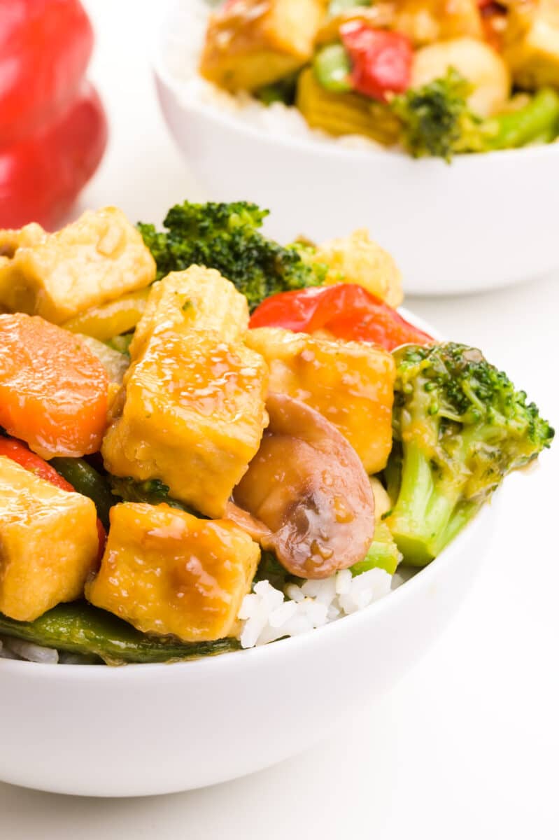 A bowl of tofu broccoli stir fry features veggies, tofu and rice. It sits in front of a red bell pepper and another bowl of the stir fry.