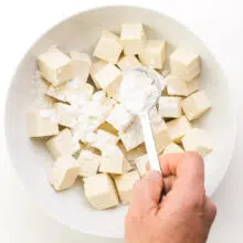 A hand holds a measuring spoon of cornstarch, sprinkling it over tofu cubes in a bowl.