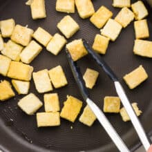A hand holds tongs, reaching them into a skillet full of tofu cubes.