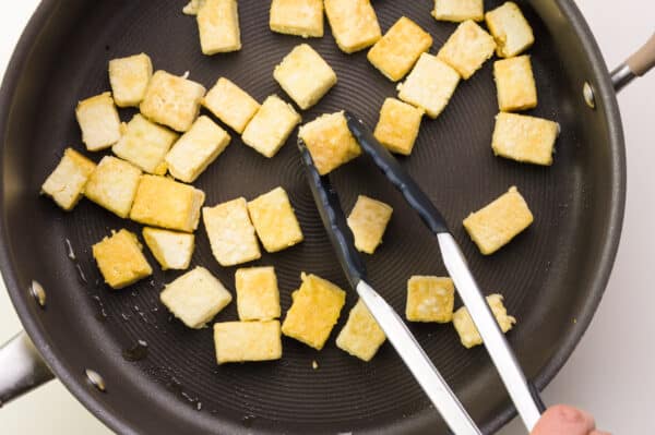 A hand holds tongs, reaching them into a skillet full of tofu cubes.