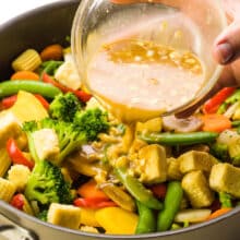 A hand holds a bowl of sauce, pouring it into a skillet full of veggies and tofu.