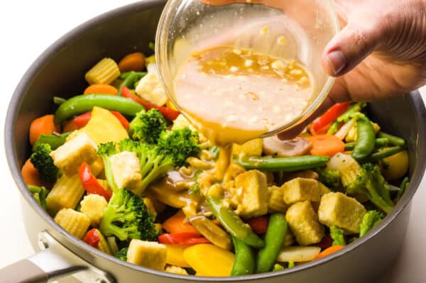 A hand holds a bowl of sauce, pouring it into a skillet full of veggies and tofu.