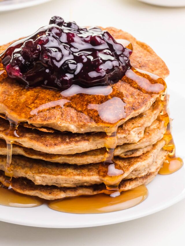 A stack of oatmeal pancakes sit on a plate with maple syrup drizzling over the sides. There is a dollop of blueberry sauce on top.