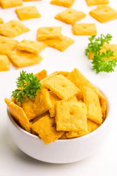 A bowl of vegan cheez-its sits in front of more of the crackers in the background. There are fresh sprigs of parsley too.
