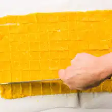 A hand holds a knife, cutting a thin orange dough into squares.