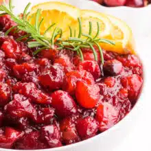 A bowl of vegan cranberry sauce sits in front of an orange and a bowl of fresh cranberries.