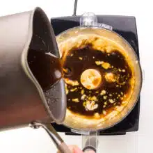 Syrup from a saucepan is being poured into a food processor with silken tofu.