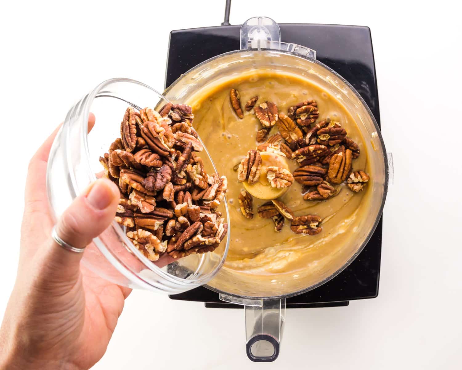 A hand holds a bowl of pecans, adding them to a food processor with a caramel-colored sauce.