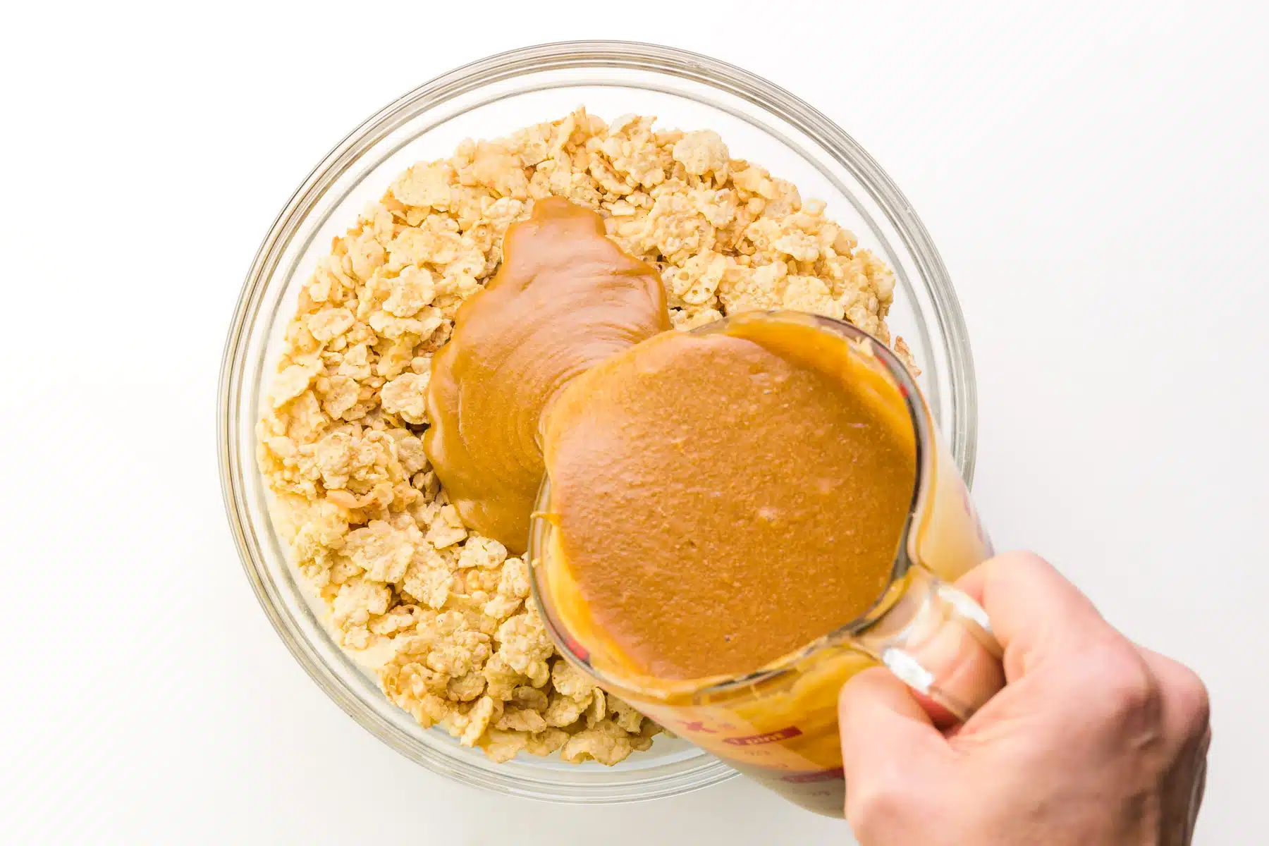 A hand holds a cup of melted peanut butter, pouring it over a bowl of cereal.