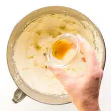 A hand holds a bowl of vanilla, pouring it into a mixing bowl with whipped butter.