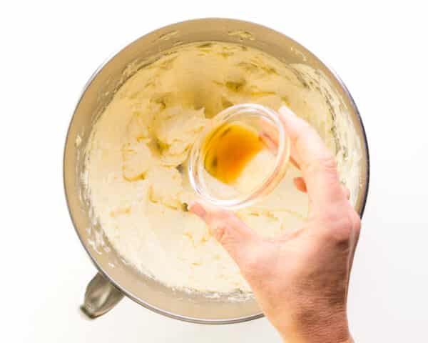 A hand holds a bowl of vanilla, pouring it into a mixing bowl with whipped butter.