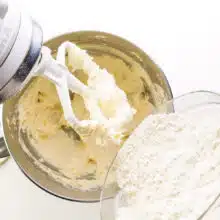 Flour is being added to a stand mixer bowl with whipped butter.