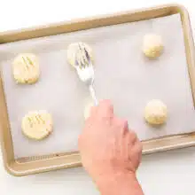 A hand holds a fork, pressing down on a cookie dough ball on a baking sheet.