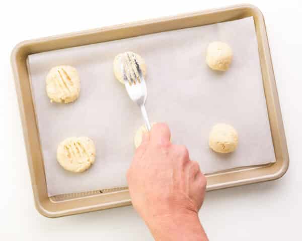 A hand holds a fork, pressing down on a cookie dough ball on a baking sheet.