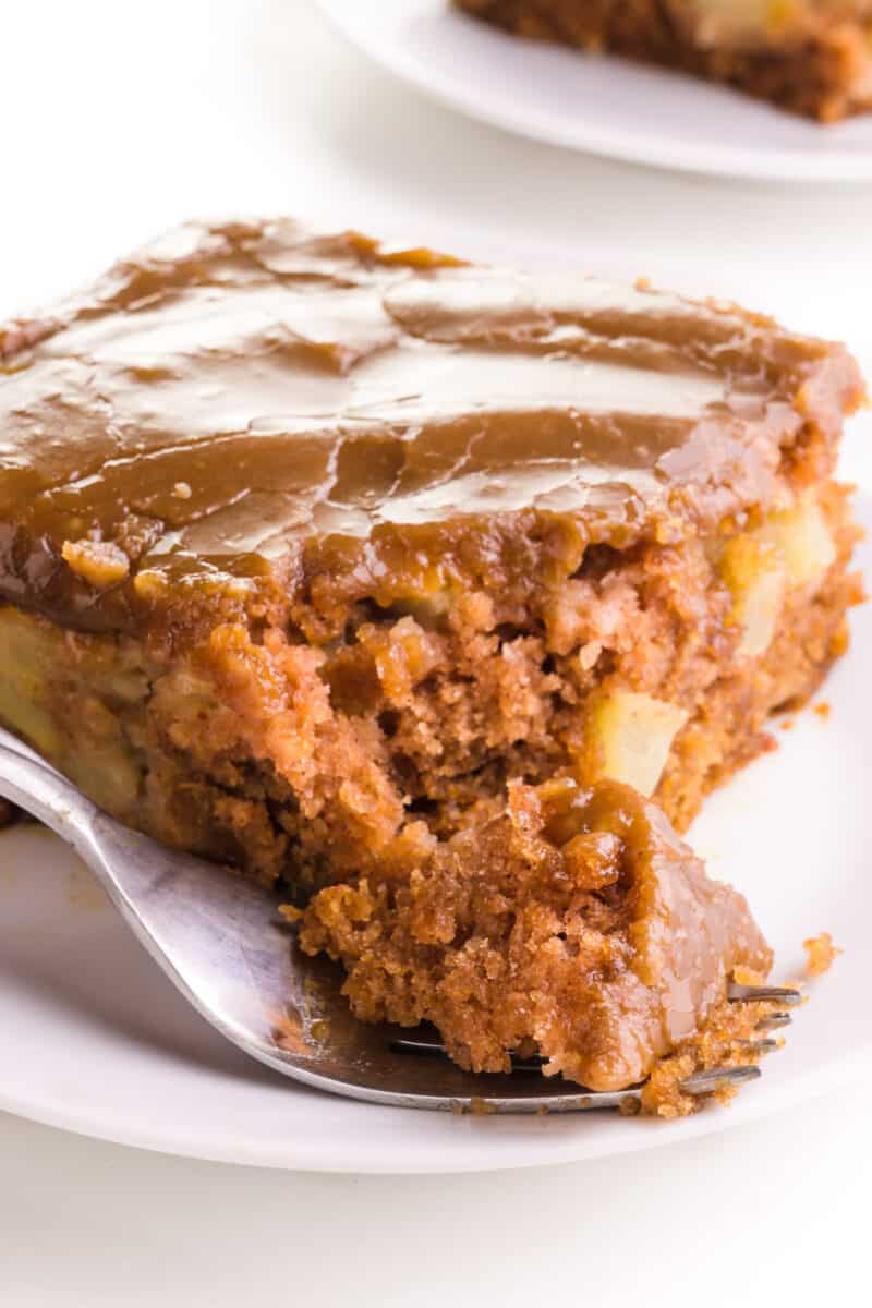 A fork holds a bite of apple cake sitting in front of the rest of the cake.