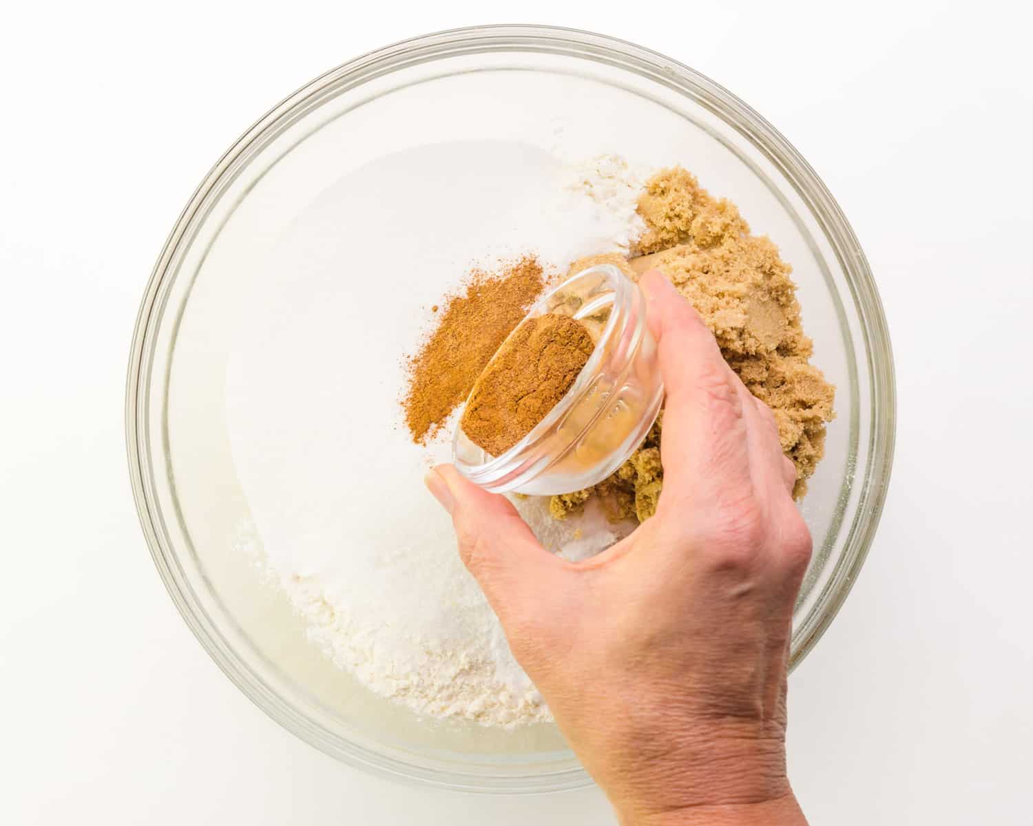 A hand pours a bowl of spices into a mixing bowl with four, sugar and other ingredients.
