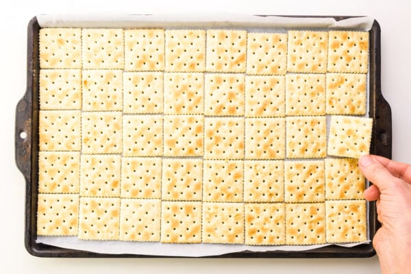 A hand places a cracker in a baking sheet lined with crackers.