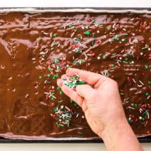 A hand spreads Christmas sprinkles over a pan with chocolate over crackers.