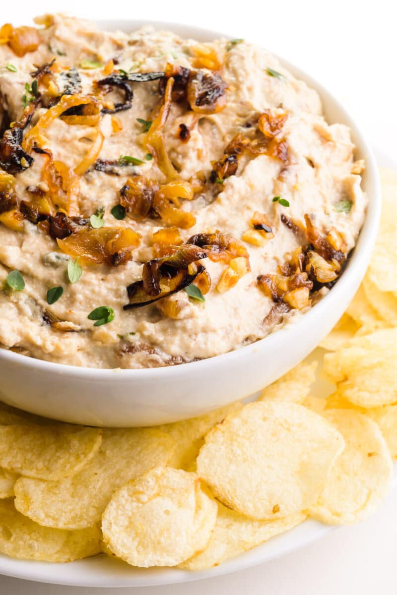 Vegan French Onion Dip sits on a plate with a bowl of potato chips.