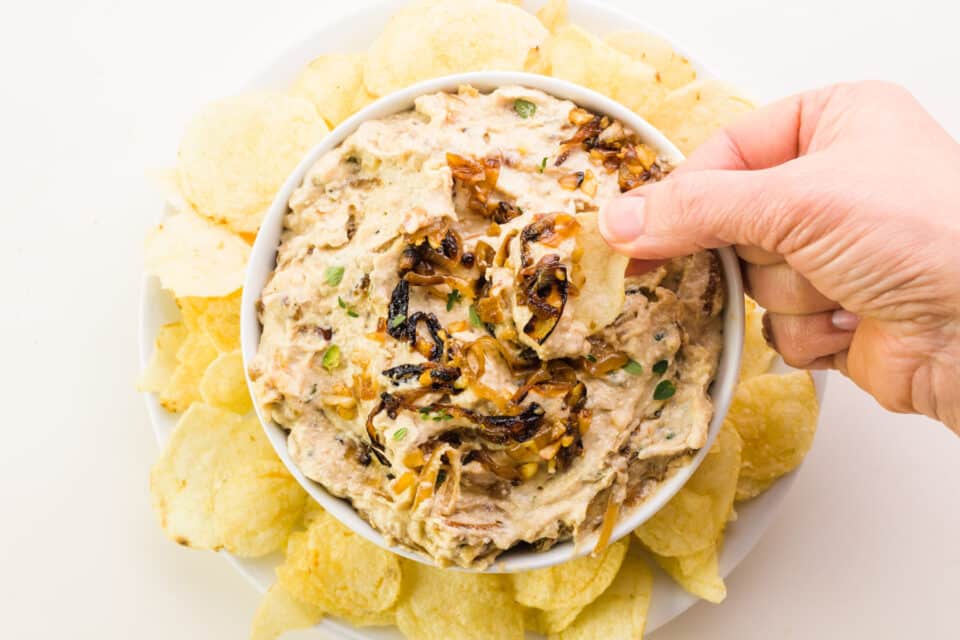 A hand holds a potato chip with dairy-free French onion dip.  It hovers over a bowl of dip surrounded by potato chips.