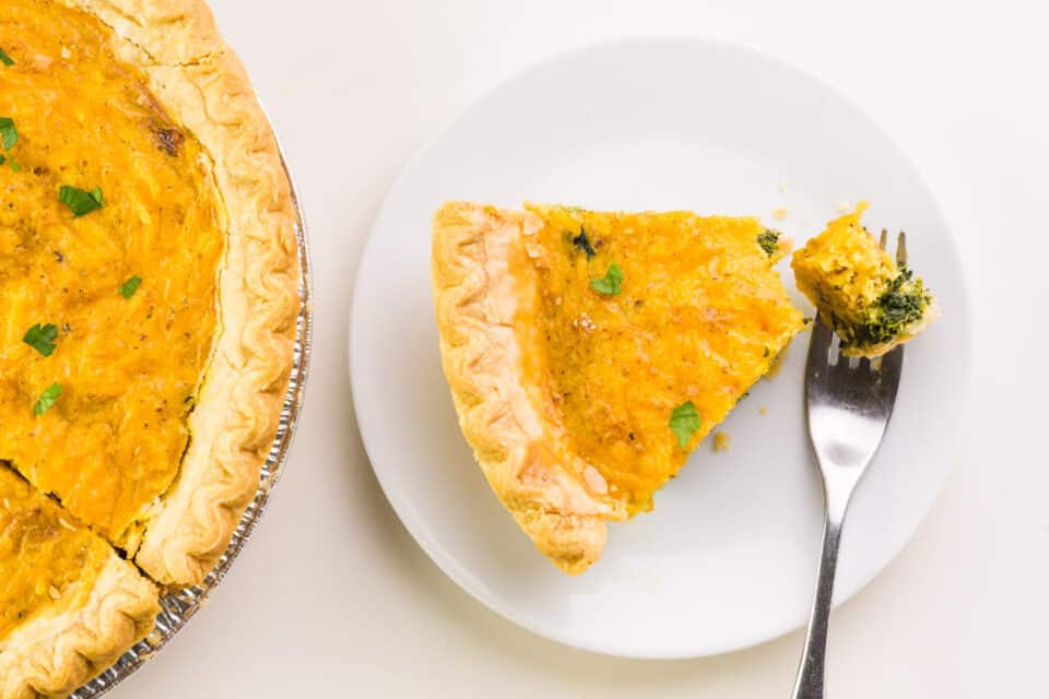 A bite out of a piece of vegan quiche sits on a plate next to the rest of the quiche in the pie pan.