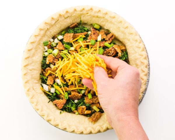 A hand spread vegan cheese across a pie crust with spinach and other ingredients.