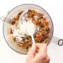 A hand holds a spoon, sprinkling flour into a food processor with toasted pecans.