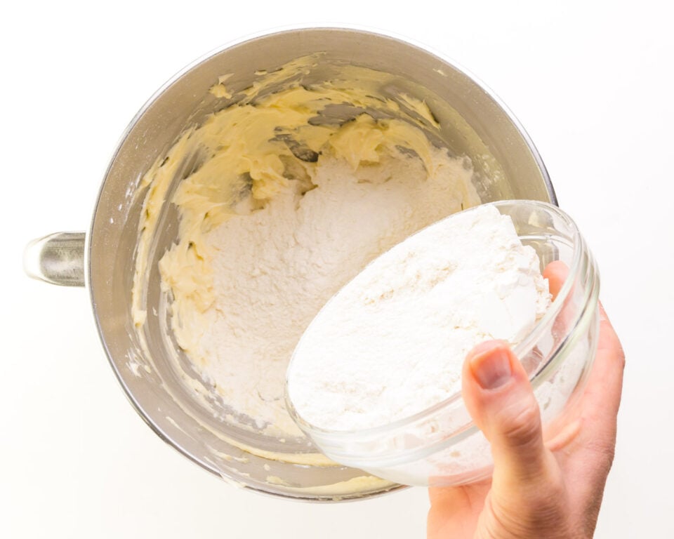 A hand holds a bowl, pouring flour into a mixing bowl with whipped butter.