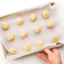 A hand holds a pan lined with parchment paper with cookie dough balls.