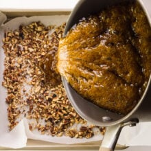 Hot sugar mixture is being poured over a pan lined with chopped nuts.