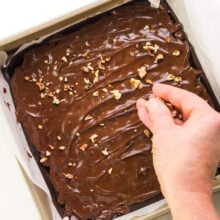 A hand spreads chopped nuts over gooey chocolate in a pan.