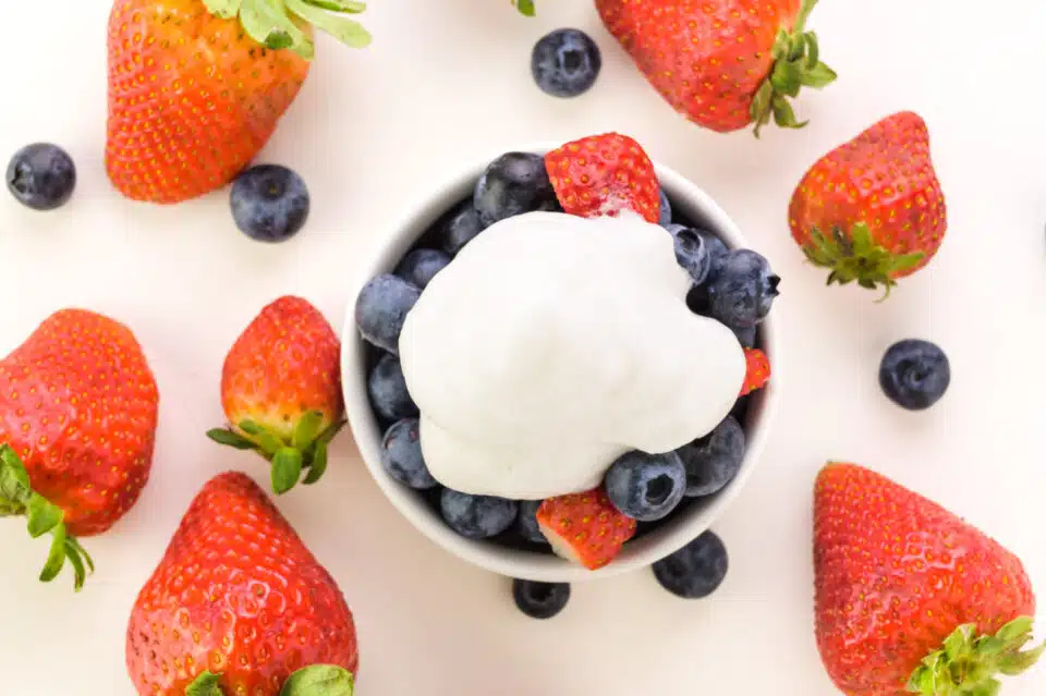 Looking down on a bowl of berries with whipped ream on top. It's surrounded by more fresh fruit.