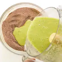 A green mixture from a food processor is being poured into a bowl with chocolate flour mixture.