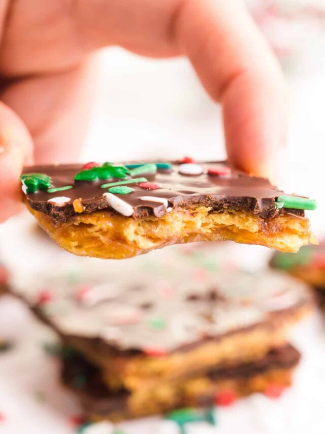A hand holds a piece of Christmas crack. It hovers over a table with another slice and sprinkles all around.