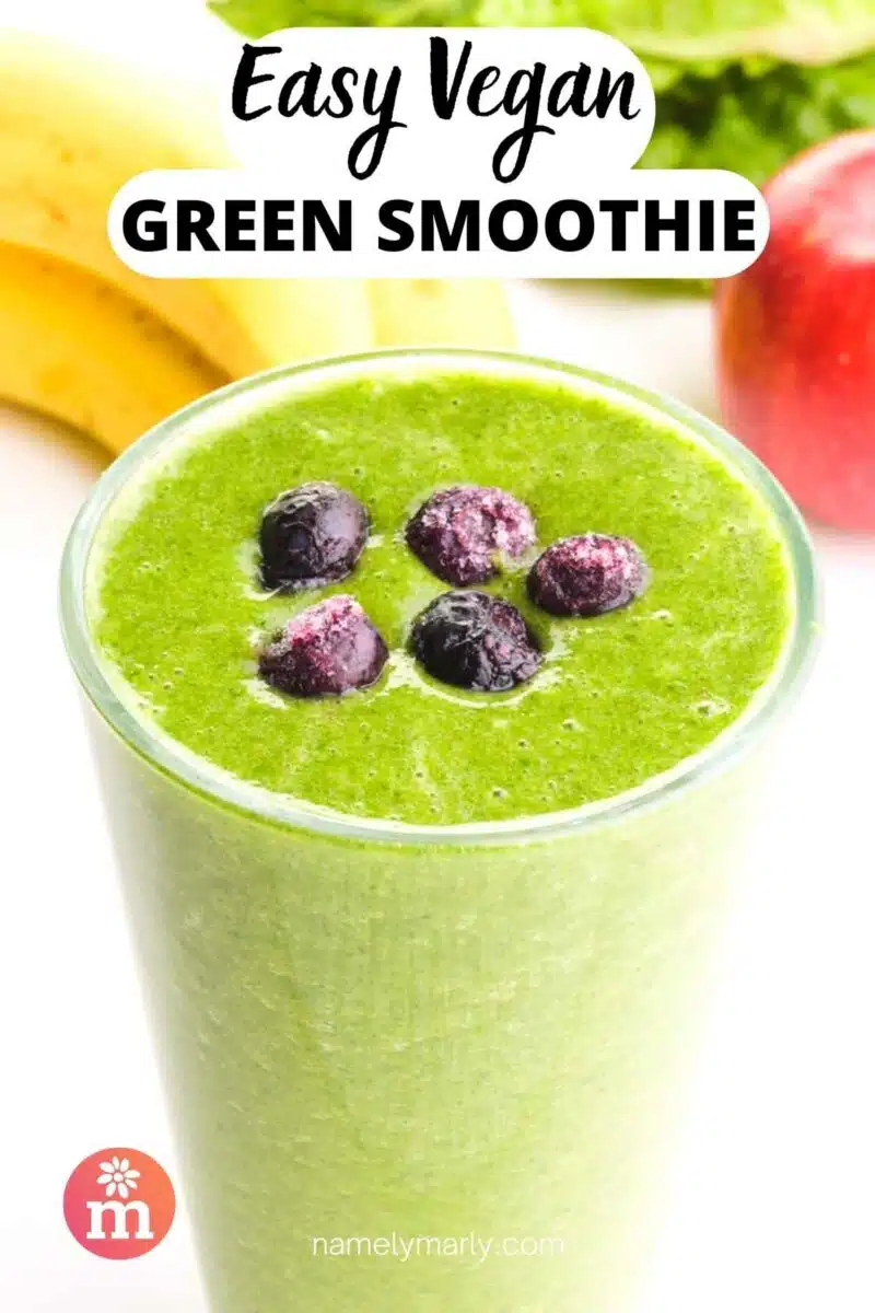 A glass of green smoothie has berries on top. There is fruit in the background. The text reads, Easy Vegan Green Smoothie.