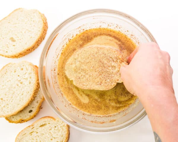 A hand places a slice of bread in a bowl, dunking it in a liquid mixture. There are more slices of bread beside the bowl.