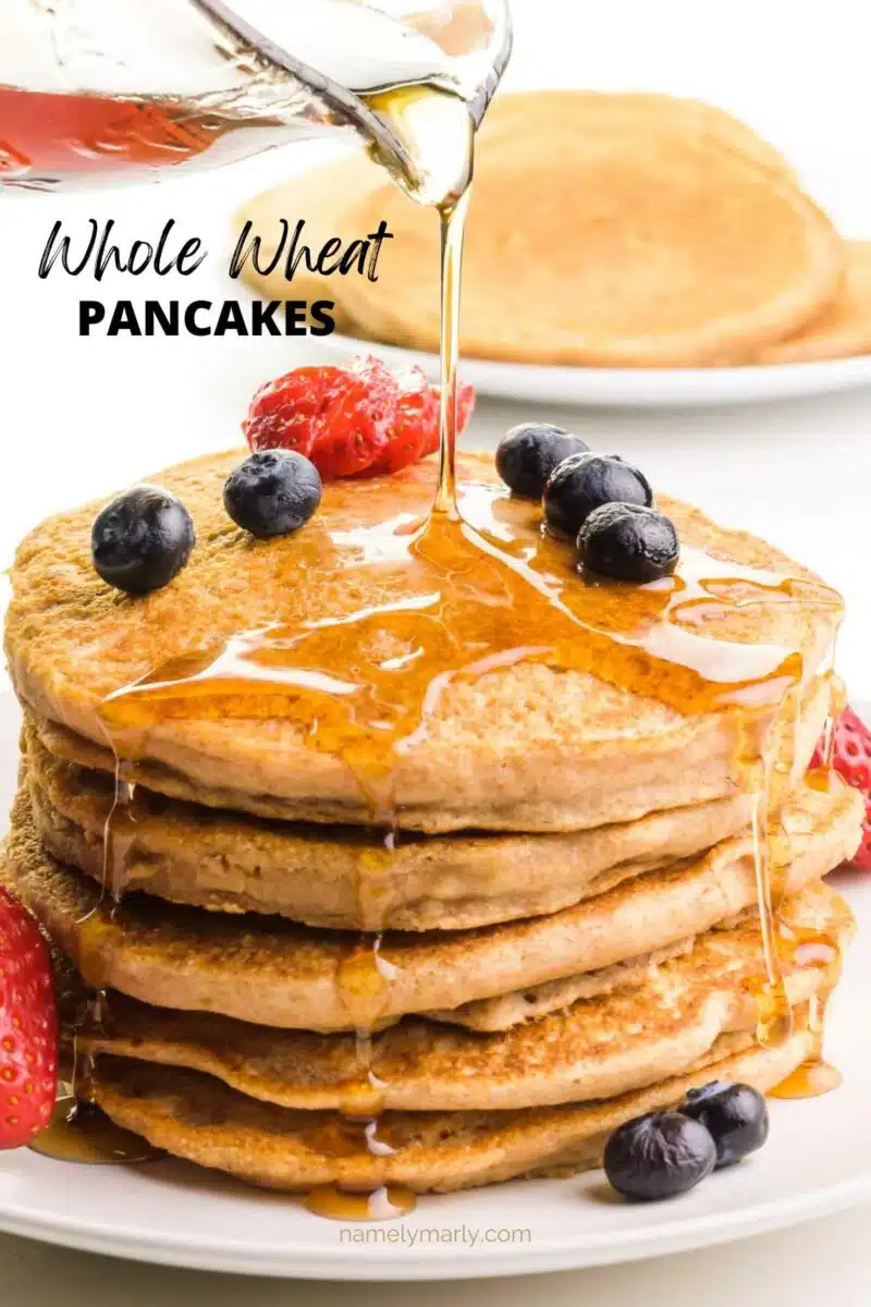 Syrup is being poured on a stack of pancakes with berries on top. There are more pancakes in the background. The text reads, Whole Wheat Pancakes.