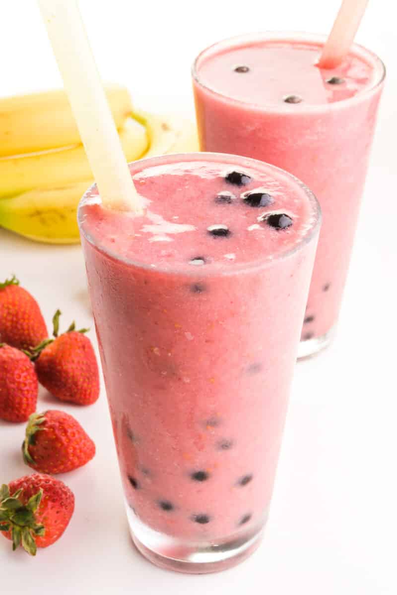 A dumb smoothie is in a glass with another behind it and fresh fruit around it.