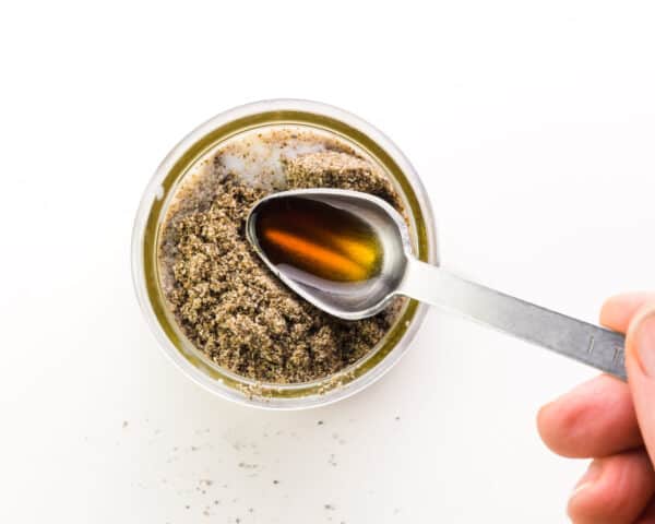 A hand holds a measuring spoon with vanilla, pouring it into a bowl with chia seeds.