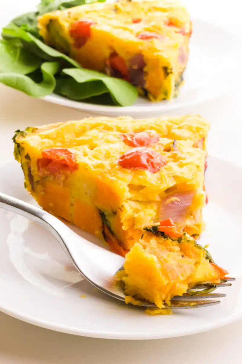 A bite of vegan frittata sits on a fork in front of the rest of the slice. There is another slice in the background and salad greens.
