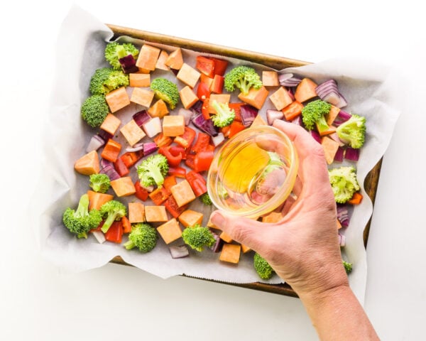 A hand pours olive oil over veggies in a sheet pan.