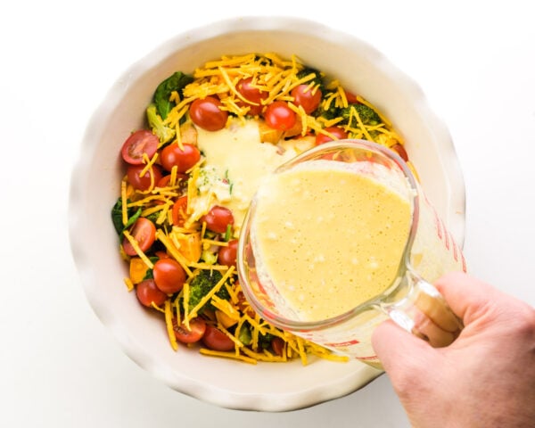 A hand pours Just Egg mixture over veggies and cheese in a pie pan.