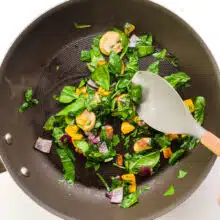 A hand holds a spatula, stirring veggies and spinach in a skillet.