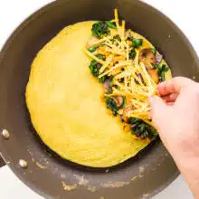 A hand distributes cheddar shreds over vegetables on one half of a whole omelet.