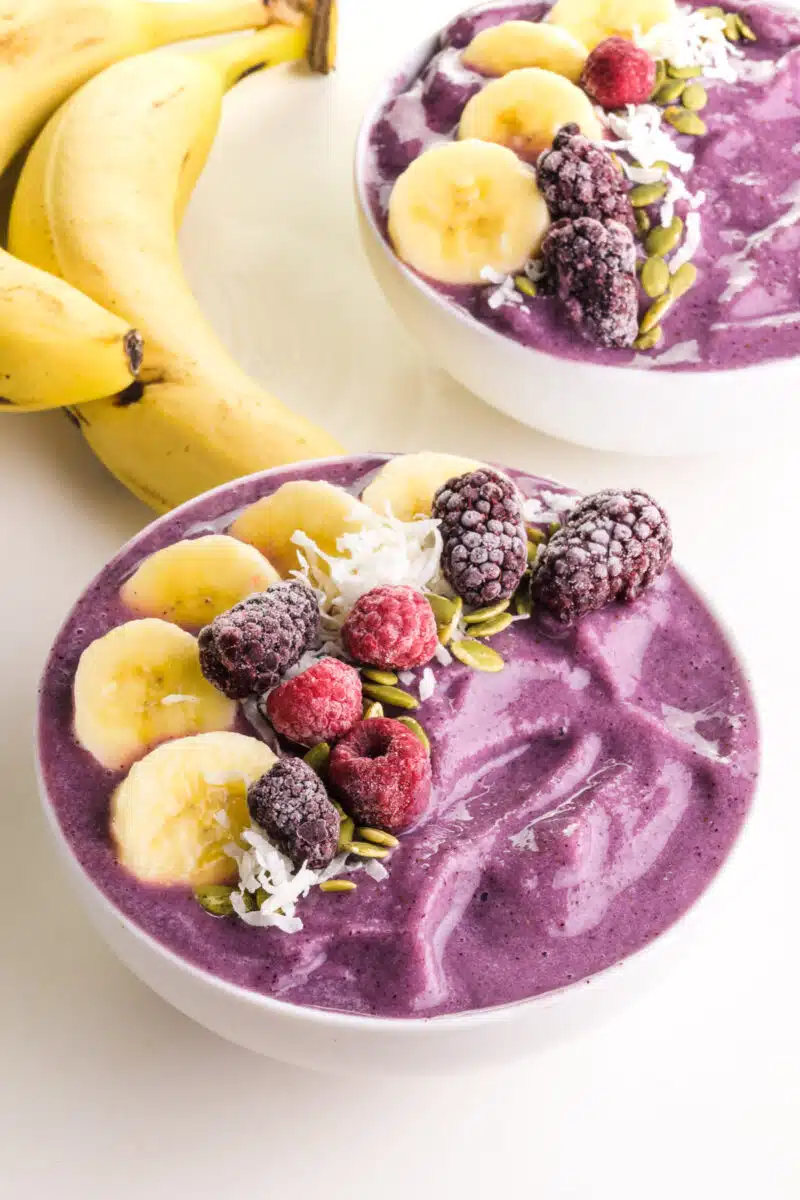 Two purple smoothie bowls have fruit on top. There are two bananas beside the bowls
