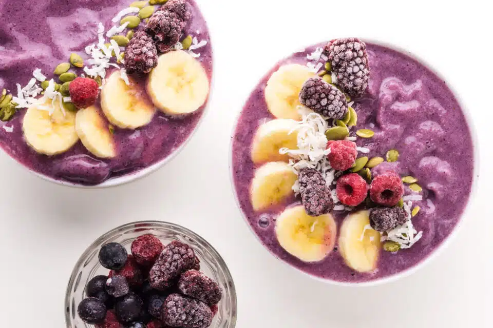 Looking down on two smoothie bowls with fresh and frozen fruit on top. There is a bowl of frozen berries nearby.