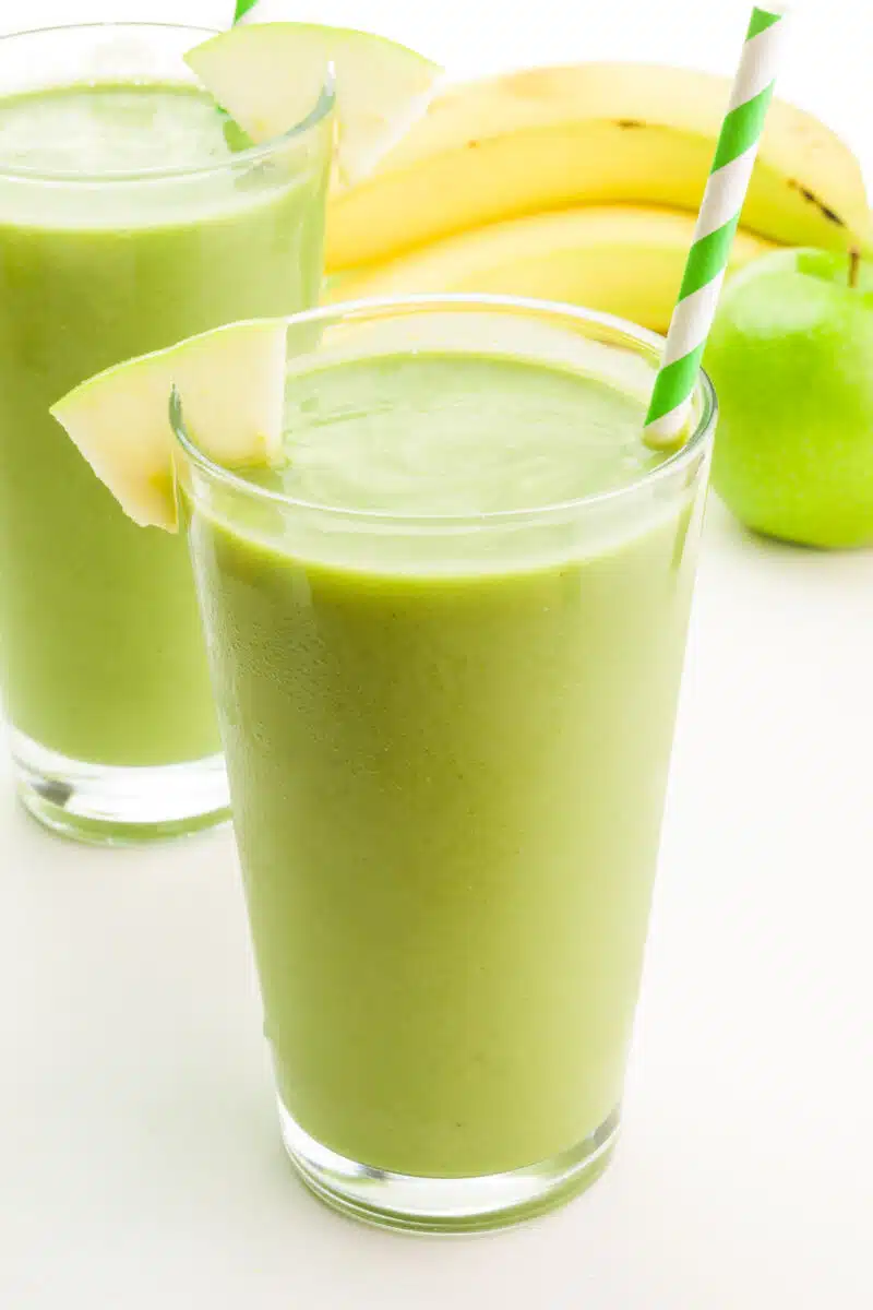A tall glass of sour apple smoothie has another glass in the background with an apple and a banana.