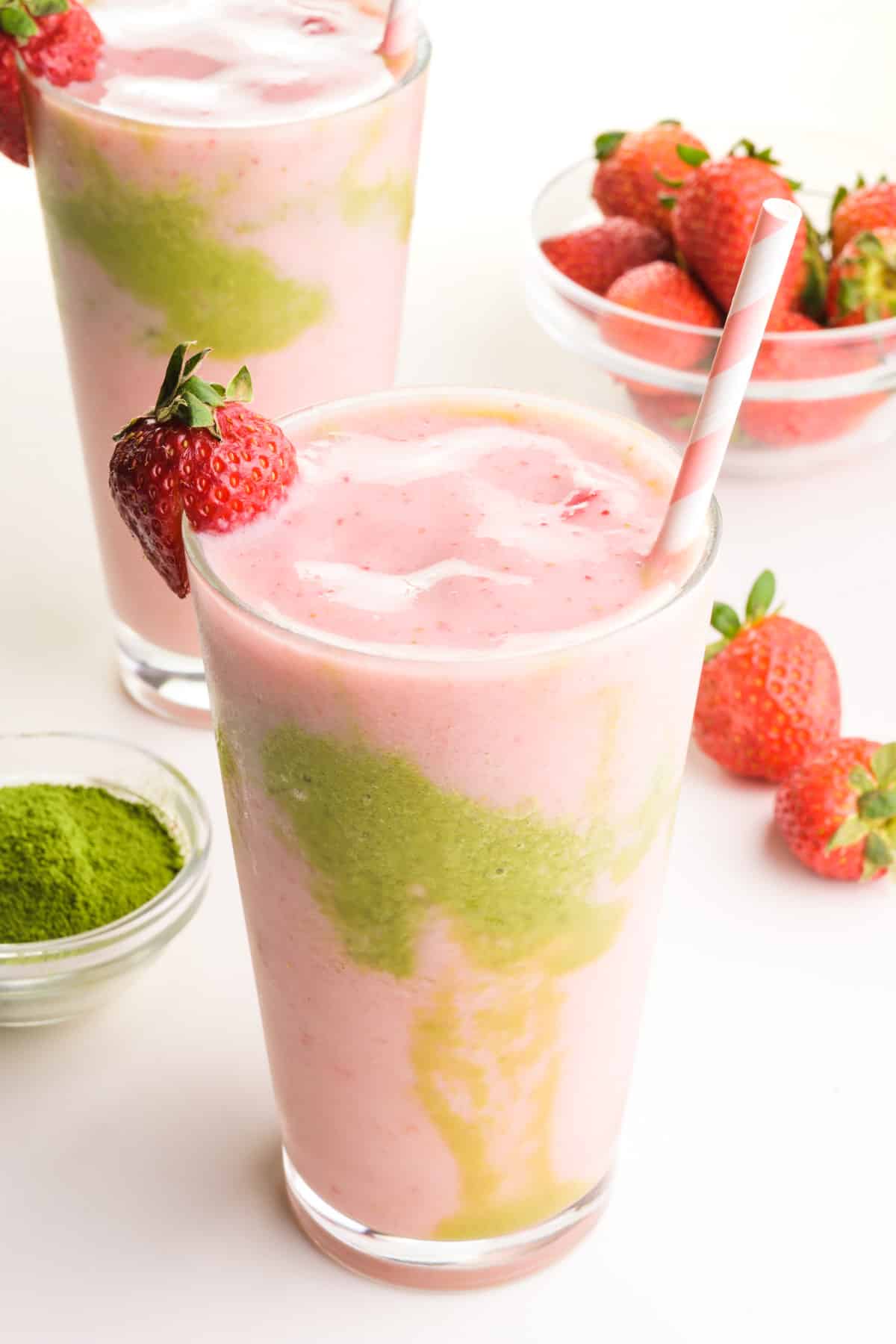 Pink and green smoothie liquid in two glasses, one in front of the other.  There is a bowl of green tea powder and strawberries around the glasses.