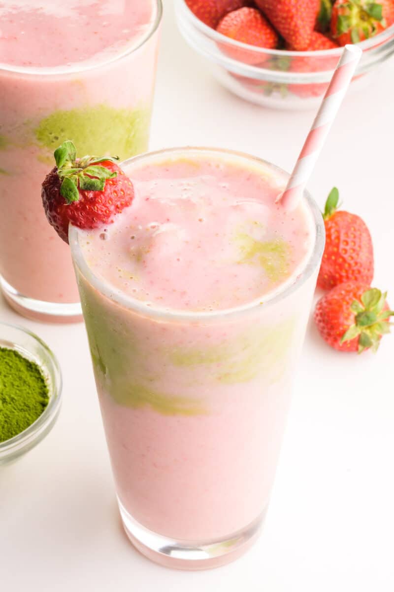 A closeup of a strawberry smoothie with matcha.  In the background is another smoothie and fresh strawberries.