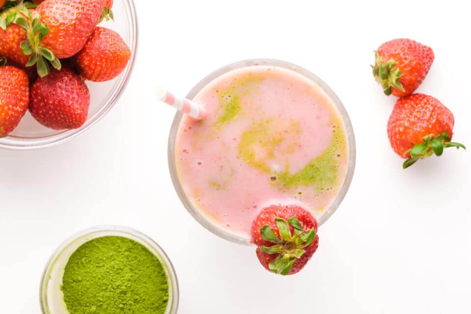 Looking at strawberry matcha smoothie in glass.  It is surrounded by fresh strawberries and matcha powder.